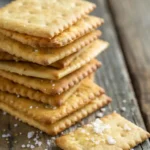 A stack of homemade gluten-free Triscuits with a golden, crispy texture and salt on top, set against a rustic wooden background.